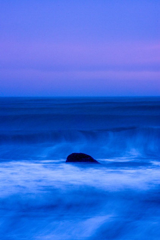 Single dark rock surrounded by silky long-exposure ocean water under a deep blue and purple twilight sky